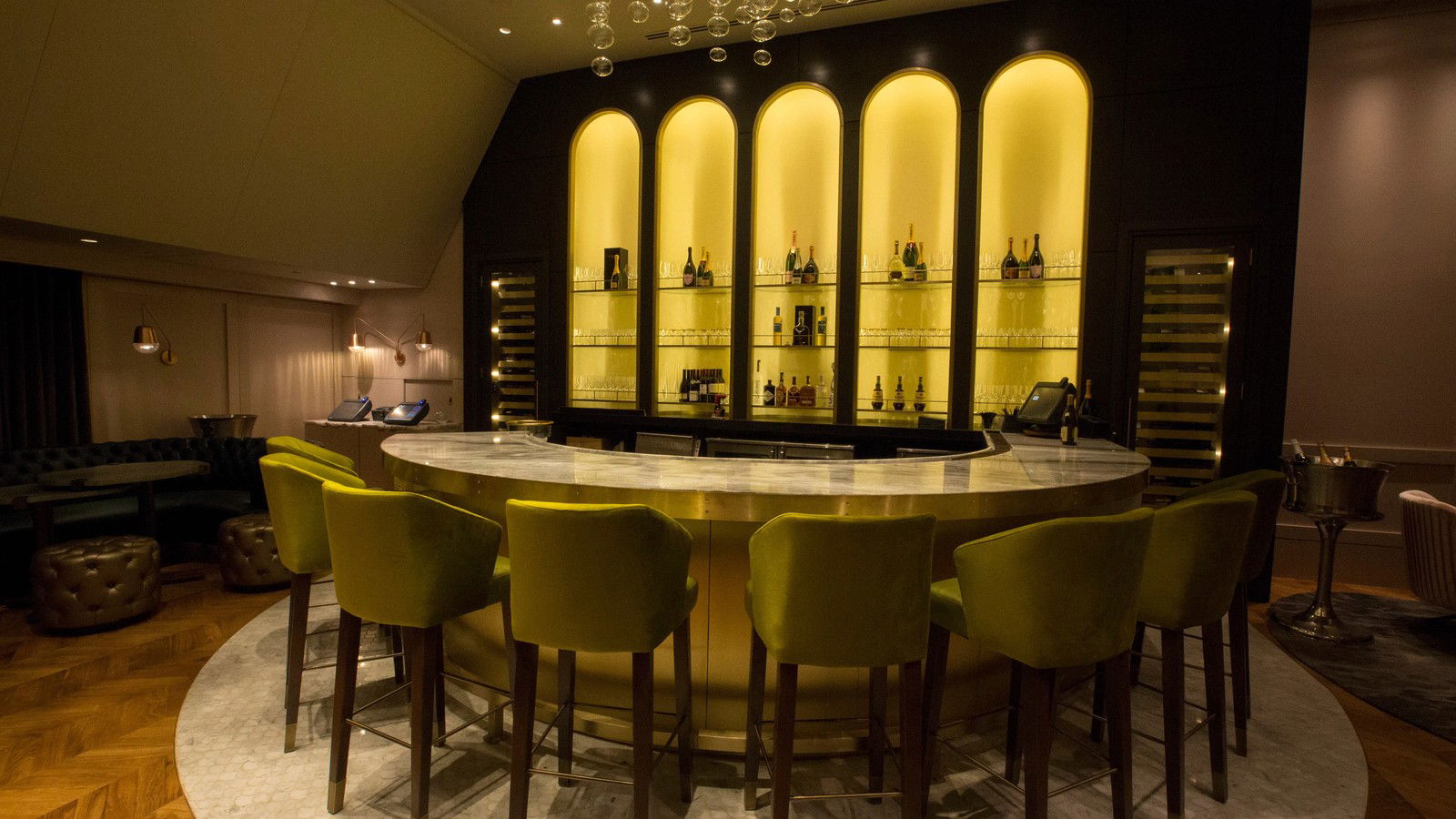 A luxurious bar with a curved marble counter and olive-green velvet stools, featuring illuminated arched shelves stocked with bottles.