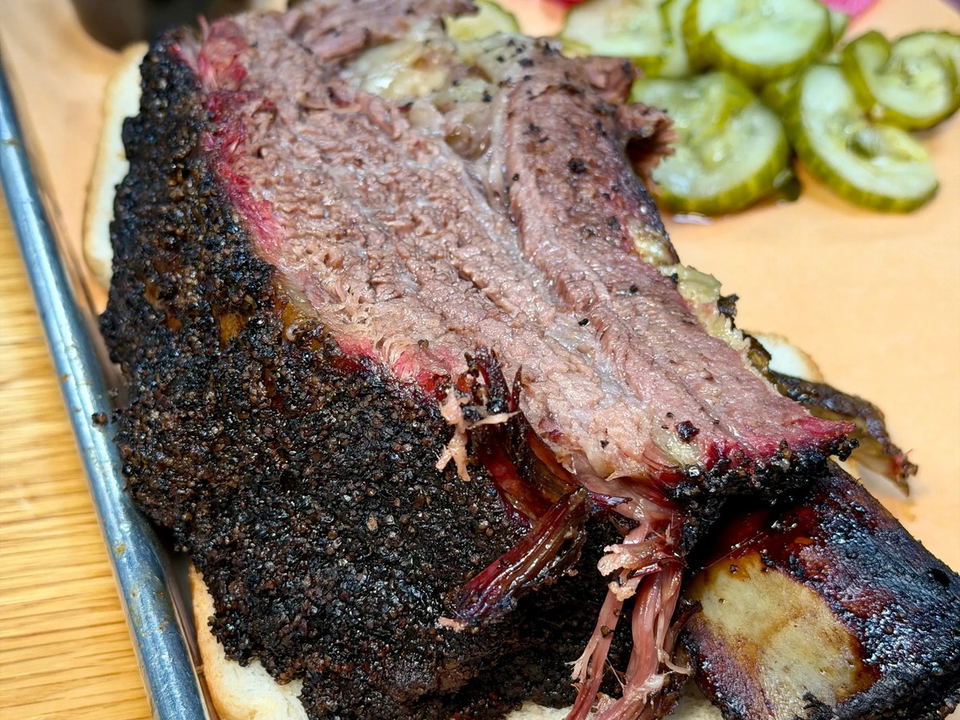A massive smoked beef rib served over white bread on a metal tray with pickled red onions, pickle slices, and a cup of BBQ sauce.