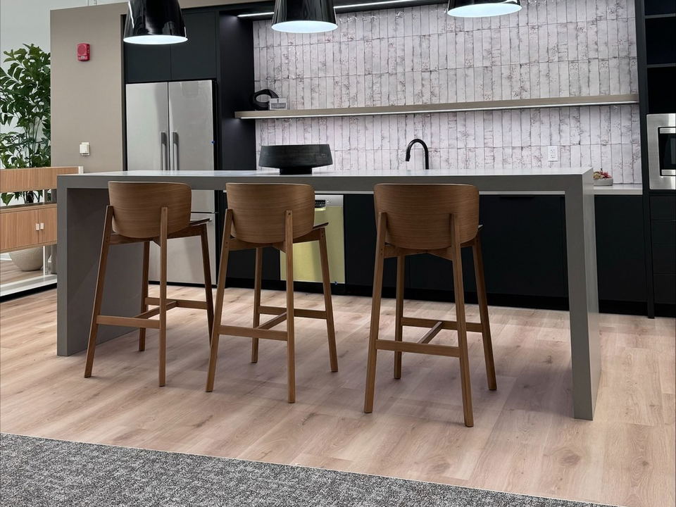 A modern office kitchen breakroom with a gray island counter, three wooden bar stools, black pendant lights, and light wood flooring.