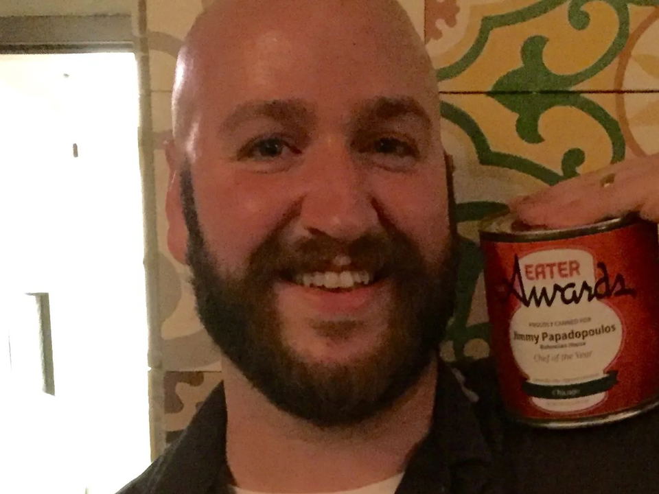 A smiling bald man with a beard wearing a "BoHo Bohemian House" chef's shirt and apron, holding a red tin can labeled "Eater Awards" with text