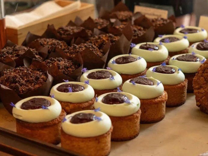 A bakery display features rows of chocolate muffins in brown paper wrappers and frosted cupcakes topped with jam and small blue flowers.
