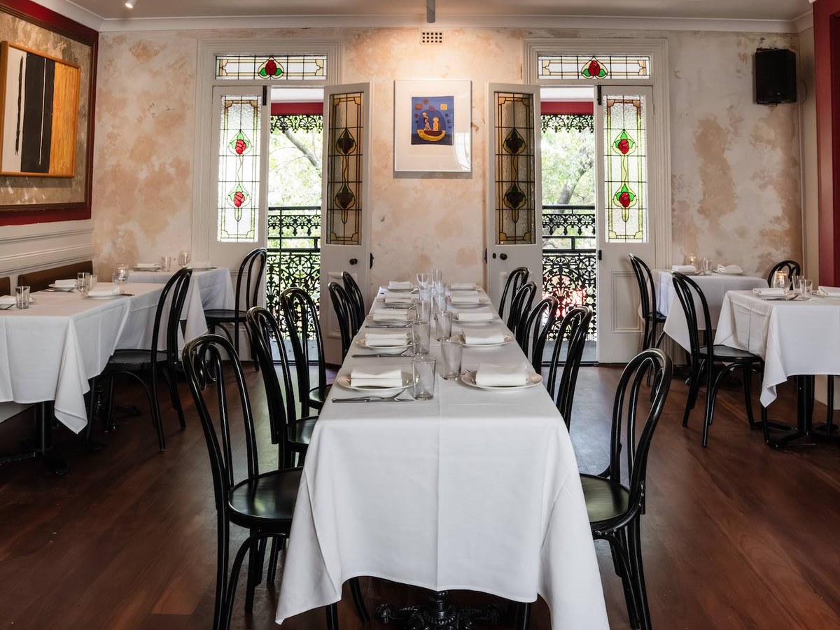 An elegantly set dining room with white-tableclothed tables, black bentwood chairs, stained-glass windows, and distressed plaster walls.