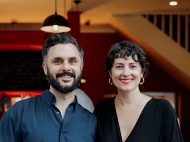 A man with a beard and a woman with curly hair smile at the camera, standing in front of a red-walled restaurant entrance.