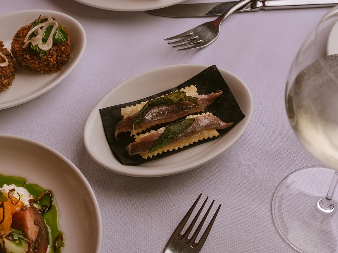 An oval white plate holds gnocco fritto topped with anchovy fillets and fried sage leaves on a dark sheet.