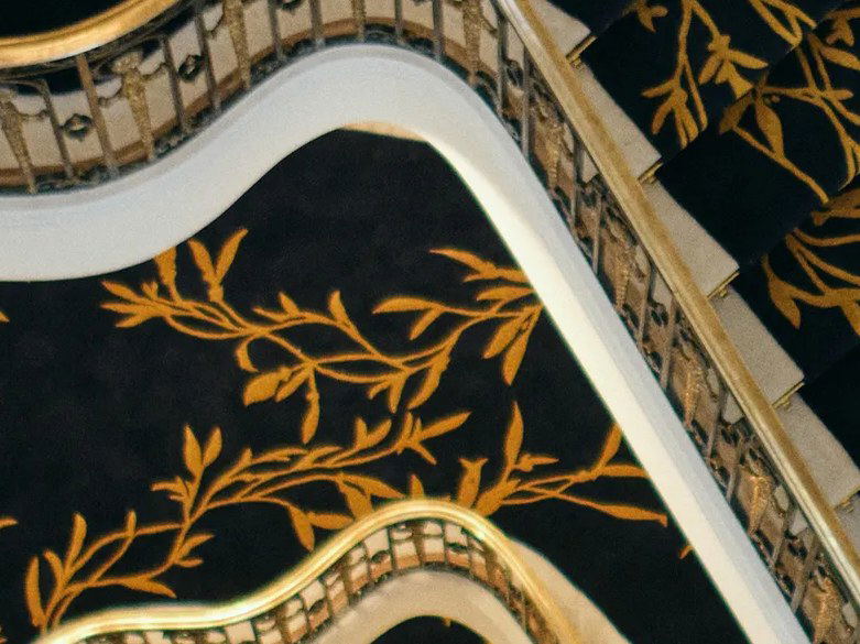 An ornate spiral staircase with black and gold botanical patterned carpet, white marble steps, and gold-trimmed railings, viewed from above.