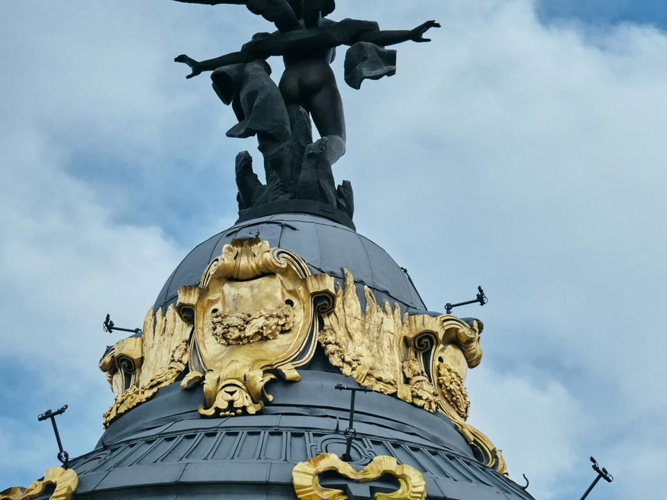 A low-angle shot of the Metrópolis Building's dome, featuring the Winged Victory sculpture with outstretched arms and wings against a cloudy sky,