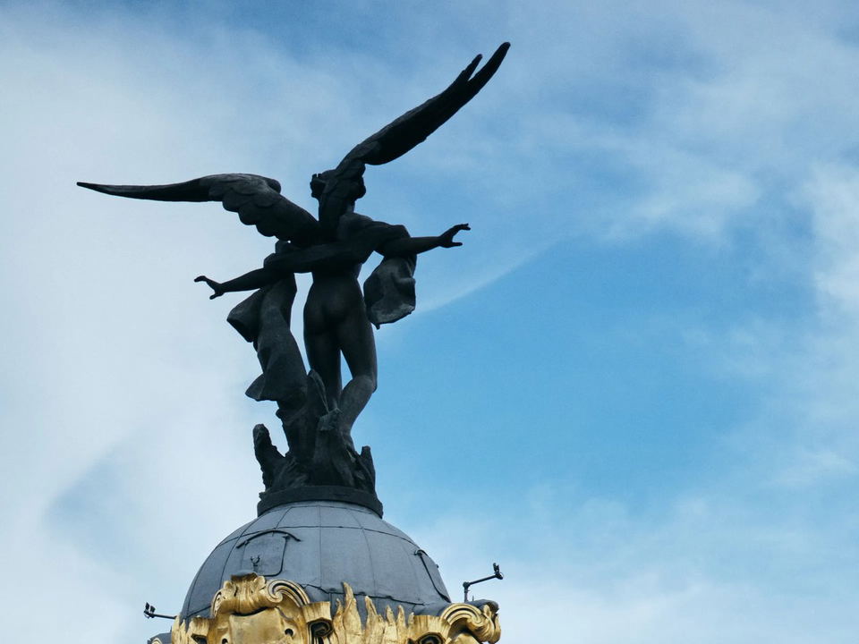 A bronze sculpture of Winged Victory with outstretched arms and wings atop the domed Metrópolis Building, set against a blue sky.