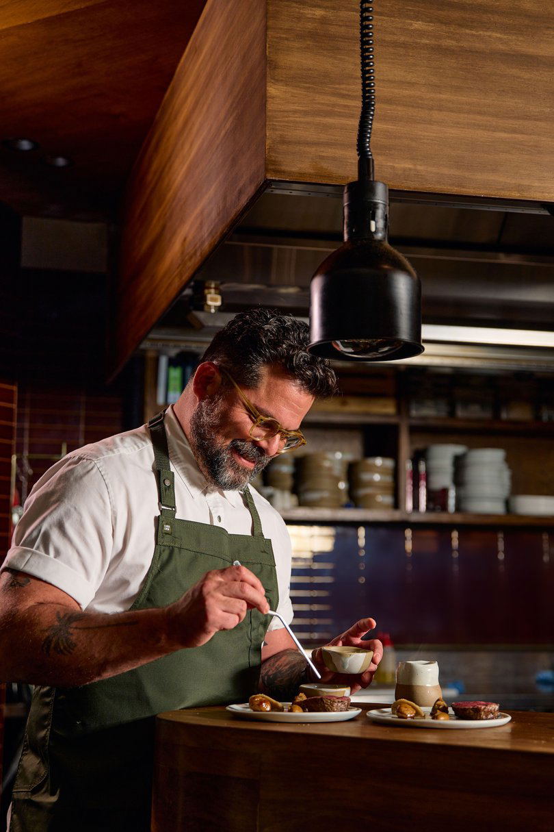 A bearded chef wearing glasses and a green apron plates food at a counter in a contemporary restaurant kitchen.