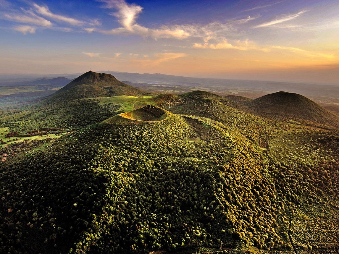 An aerial view of the Massif Central region shows several green, tree-covered volcanic cinder cones under a blue and orange sky.