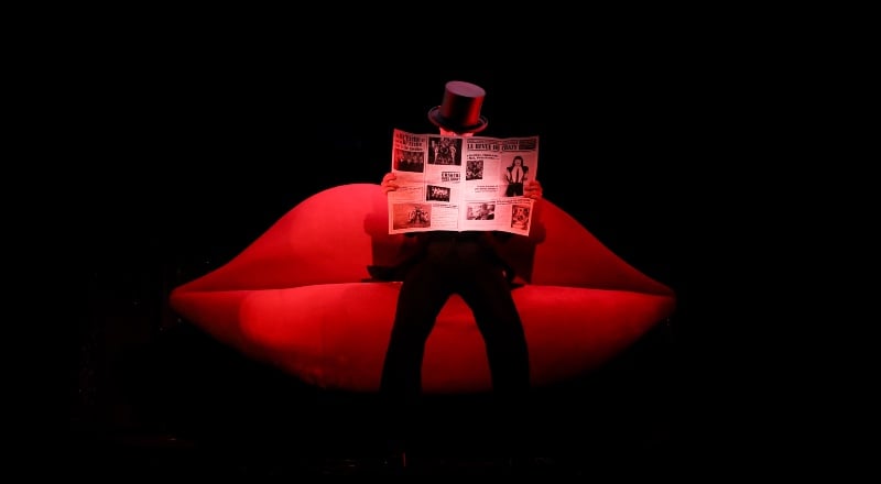 A theatrical cabaret stage setup with a large illuminated red lips sculpture and a silhouetted performer reading a newspaper