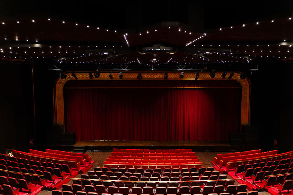 Interior view of a theater auditorium showing red velvet seats, ornate proscenium arch, red stage curtain, and house lights