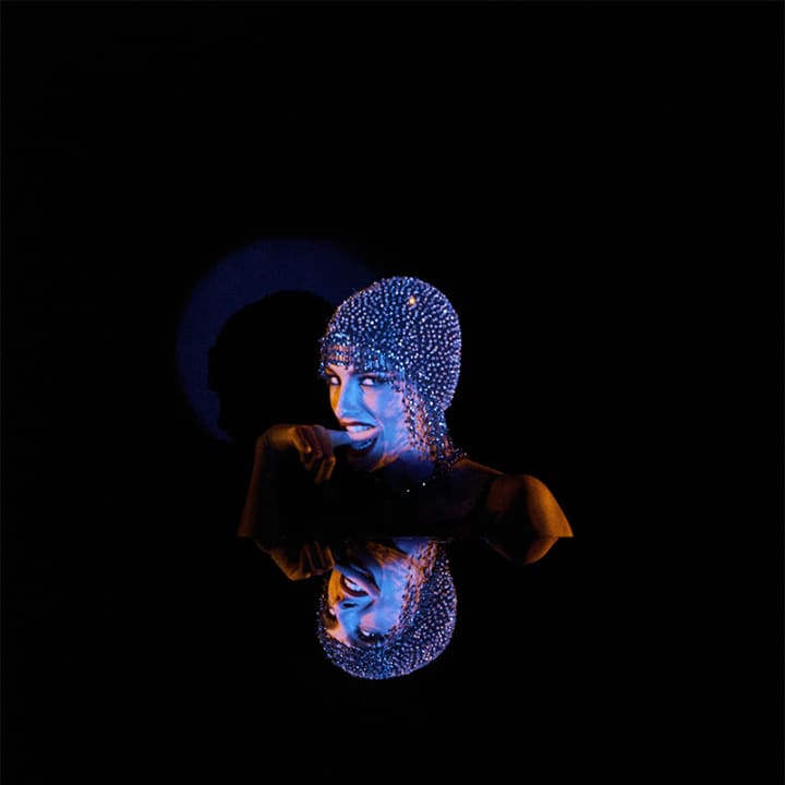 A performer in elaborate costume with studded/beaded headpiece and body decoration under dramatic blue and orange stage lighting against a black backg