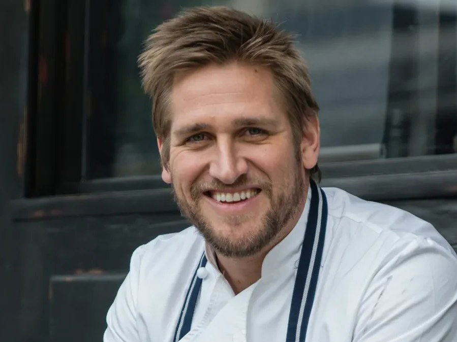 Curtis Stone, a chef in a white jacket and striped apron, sits smiling on a wooden bench outside a dark building.