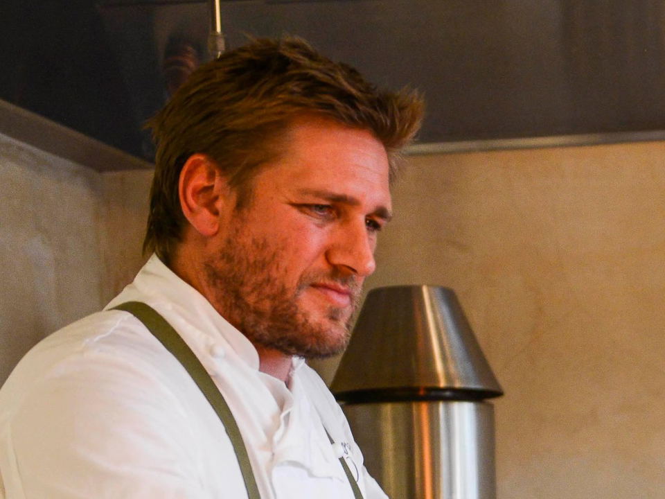 A man with styled brown hair and facial hair wearing a white chef's coat and olive-colored apron, photographed indoors in profile view.