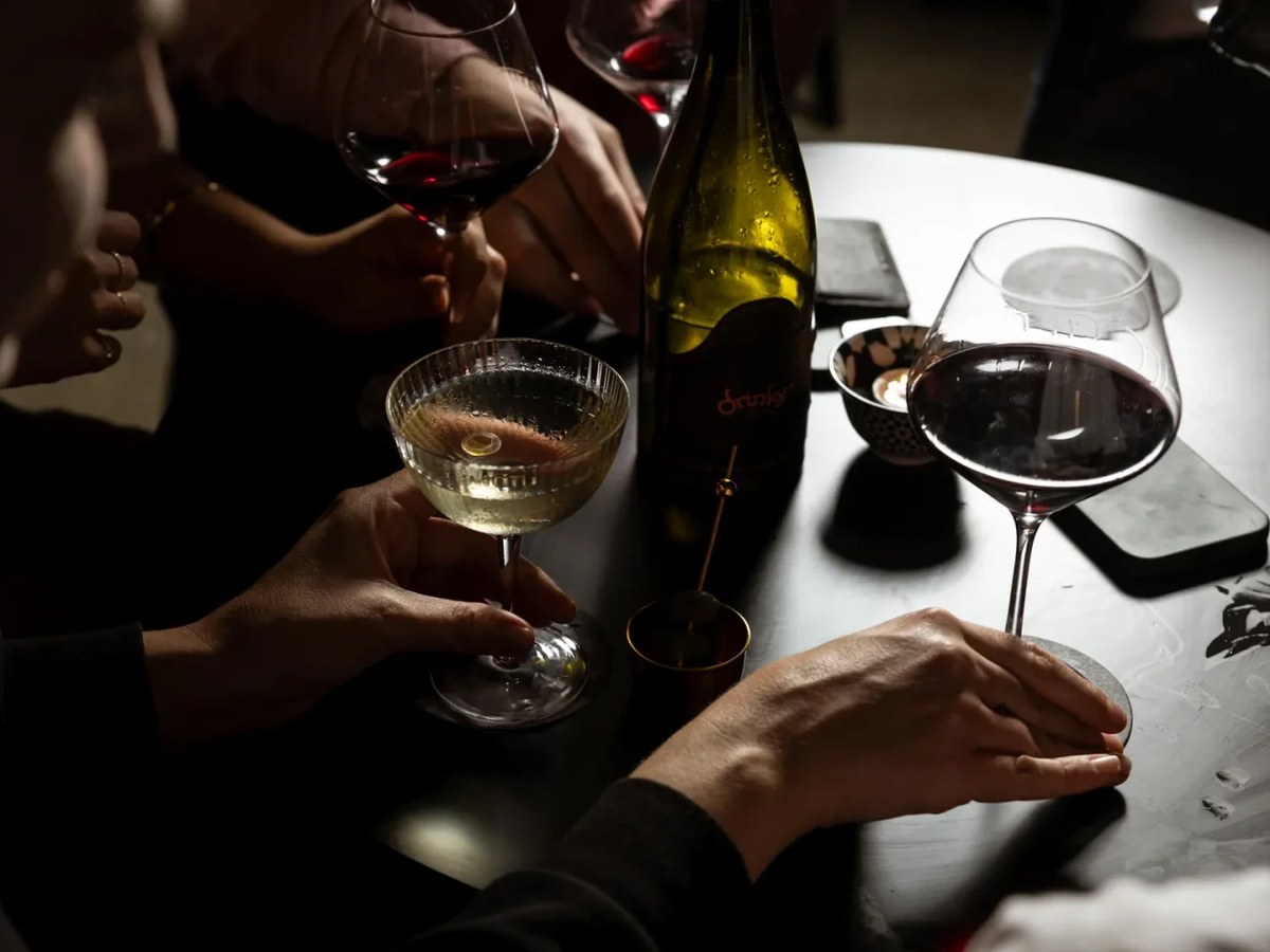 Close-up overhead shot of people at a dark bar table holding wine glasses and a coupe cocktail glass, with a green wine bottle (partially readable lab