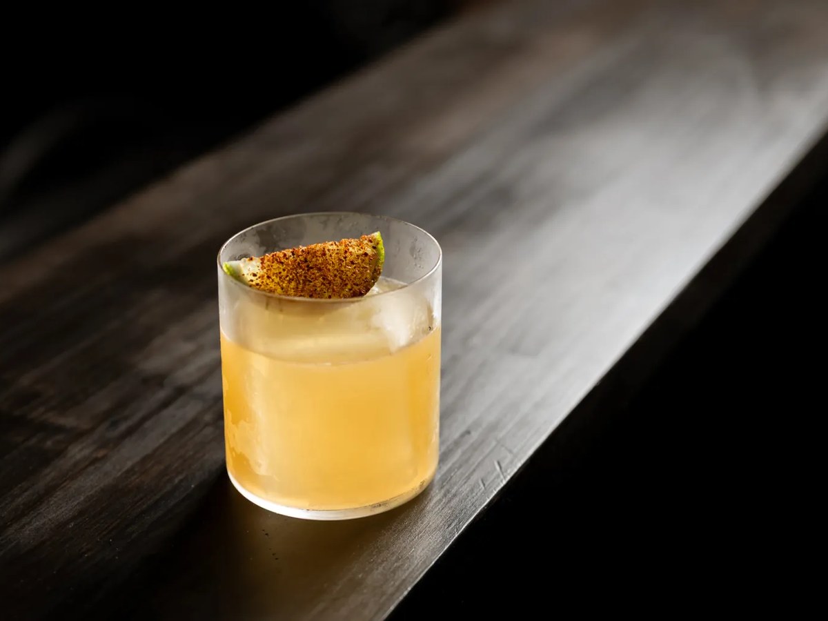 A finished cocktail in a rocks glass with a large ice cube and a chili-dusted lime wedge garnish, resting on a dark wooden bar top, consistent with th