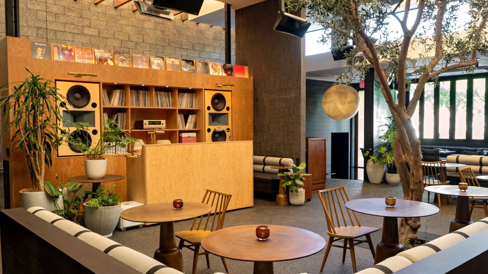 A modern restaurant interior with mid-century furniture, built-in hi-fi speakers in wood cabinetry, and an indoor tree.