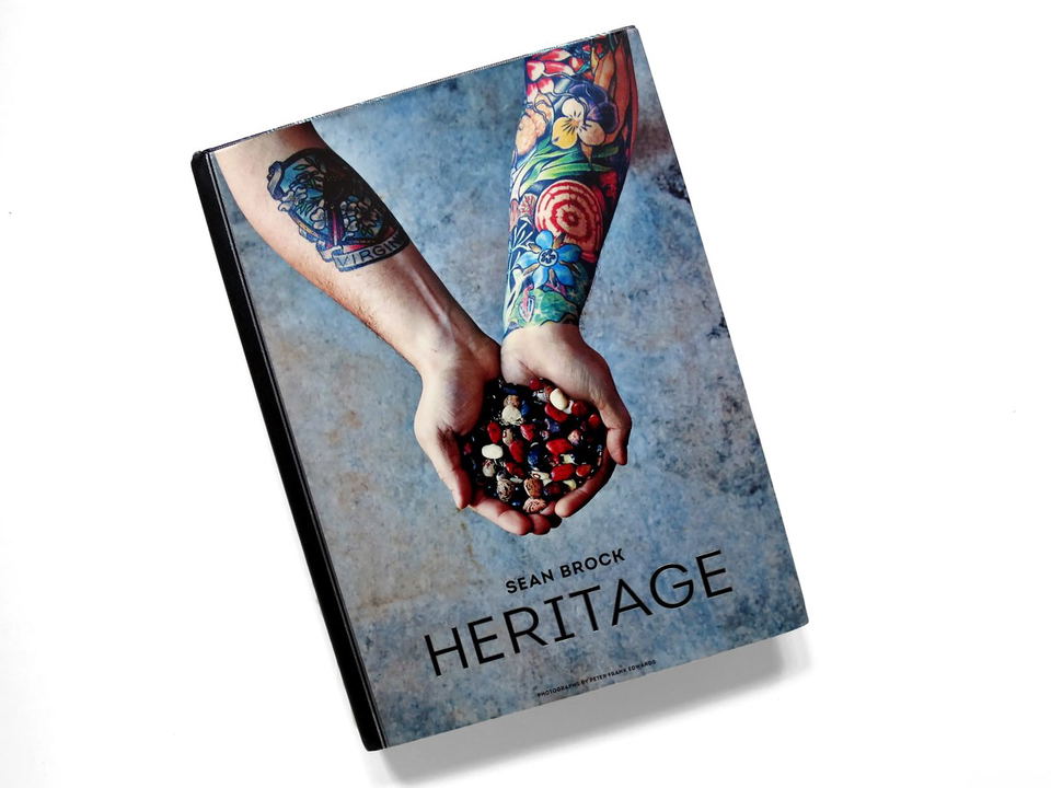 The cover of Sean Brock's Heritage cookbook, featuring two tattooed hands holding a variety of colorful beans against a textured background.