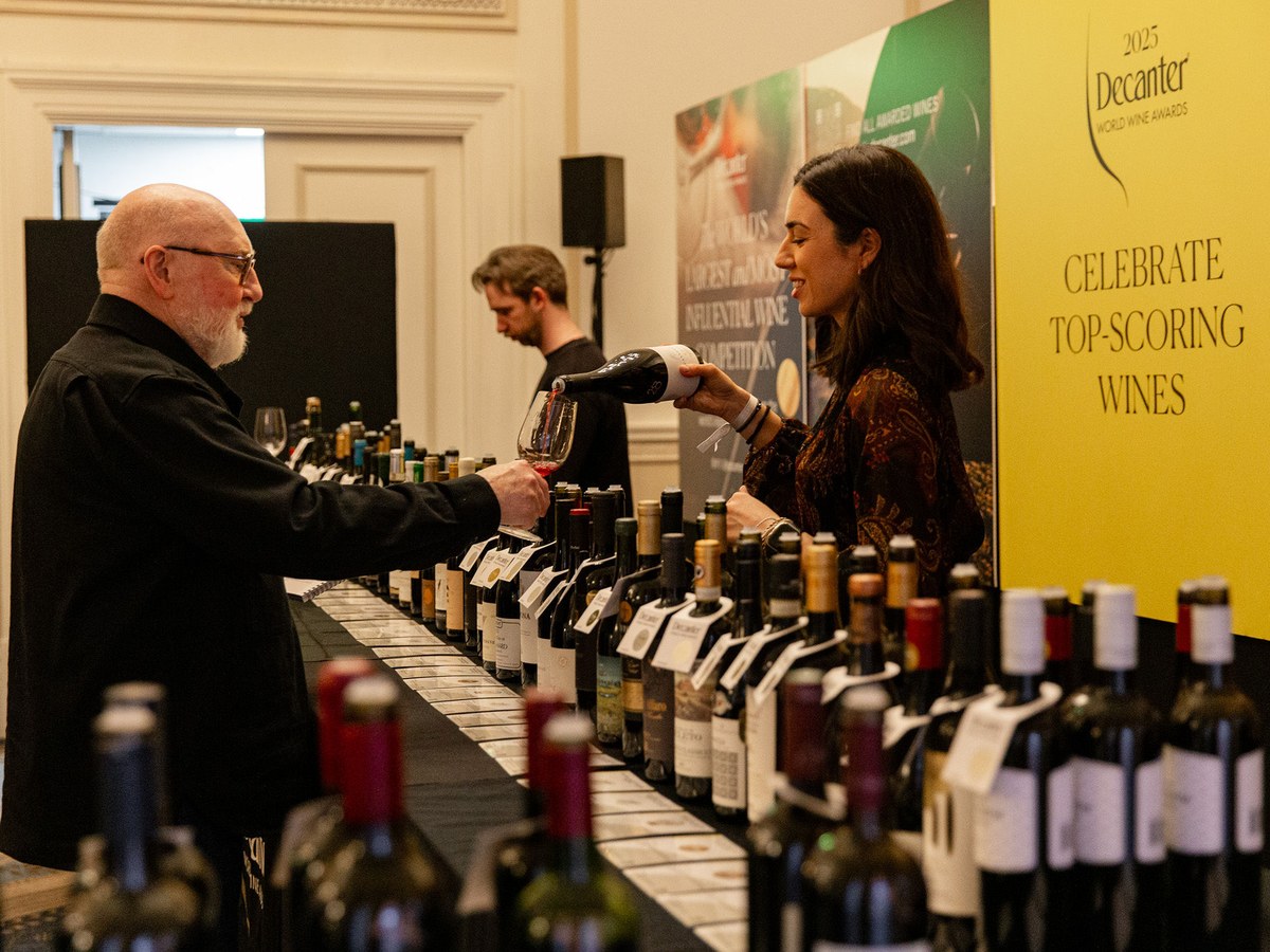 A woman pours red wine from a bottle into a glass held by a man at a long table laden with numerous wine bottles, with a yellow banner in the