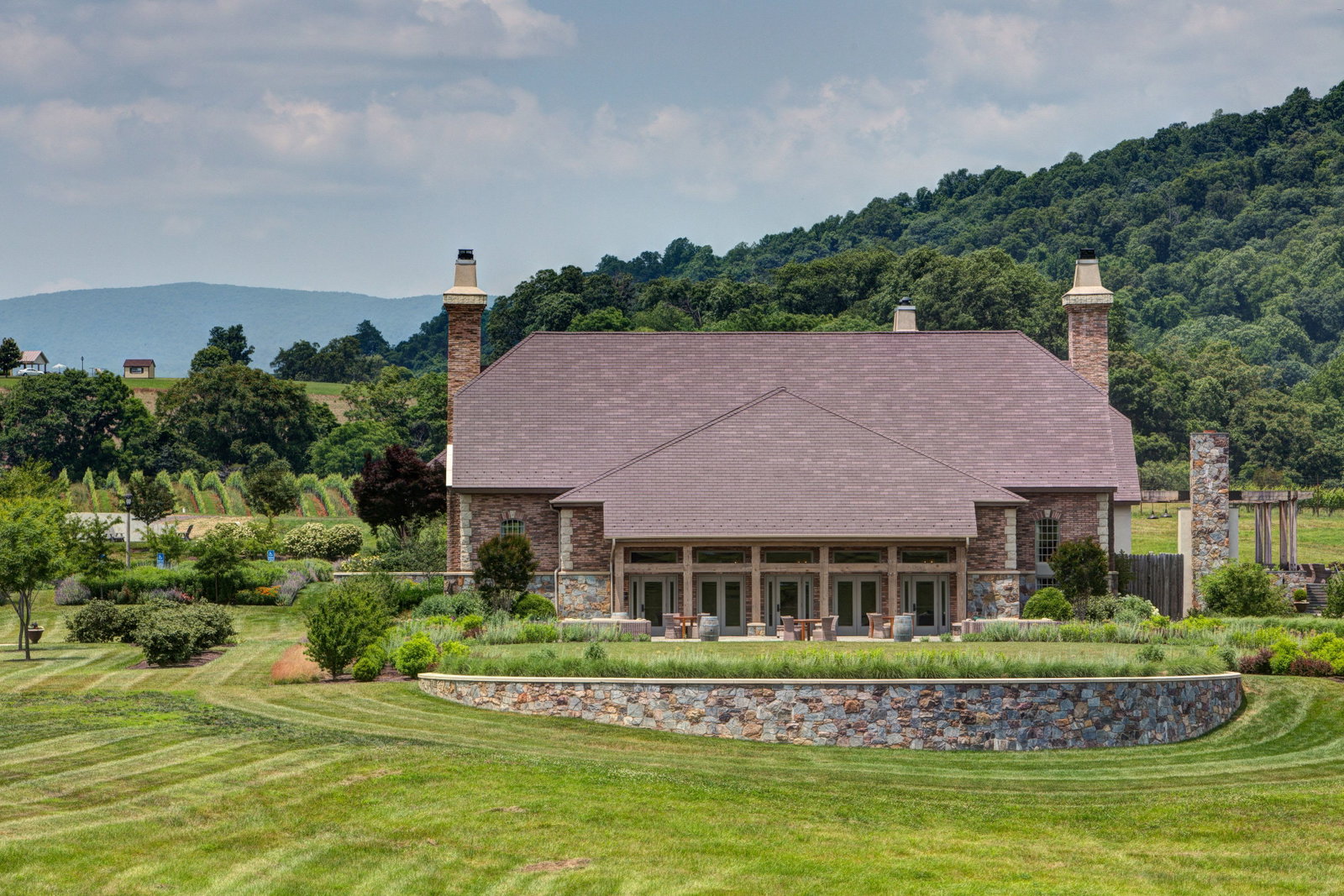 A large, elegant building with a brown roof and stone facade, flanked by two tall chimneys, sits amidst lush green lawns and manicured gardens. In