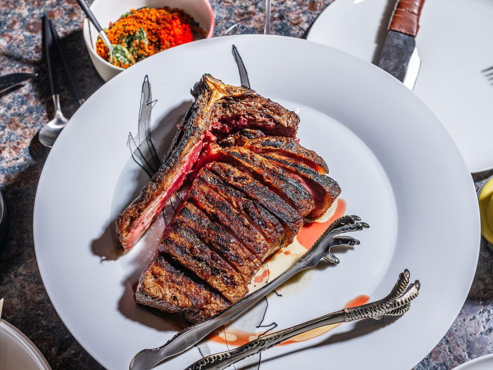 A high-end restaurant plating of a grilled bone-in meat with decorative tongs, a wine glass, and side dishes on a granite table.