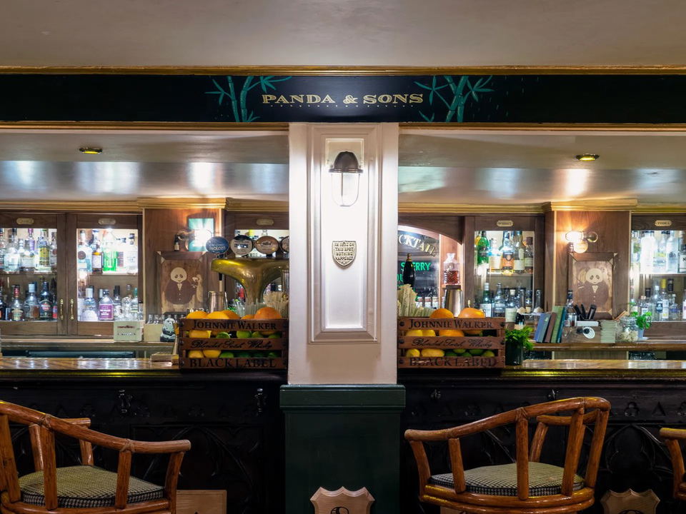 The interior of Panda & Sons bar, featuring a dark wood bar, backlit shelves filled with various liquor bottles, and a sign with bamboo motifs and