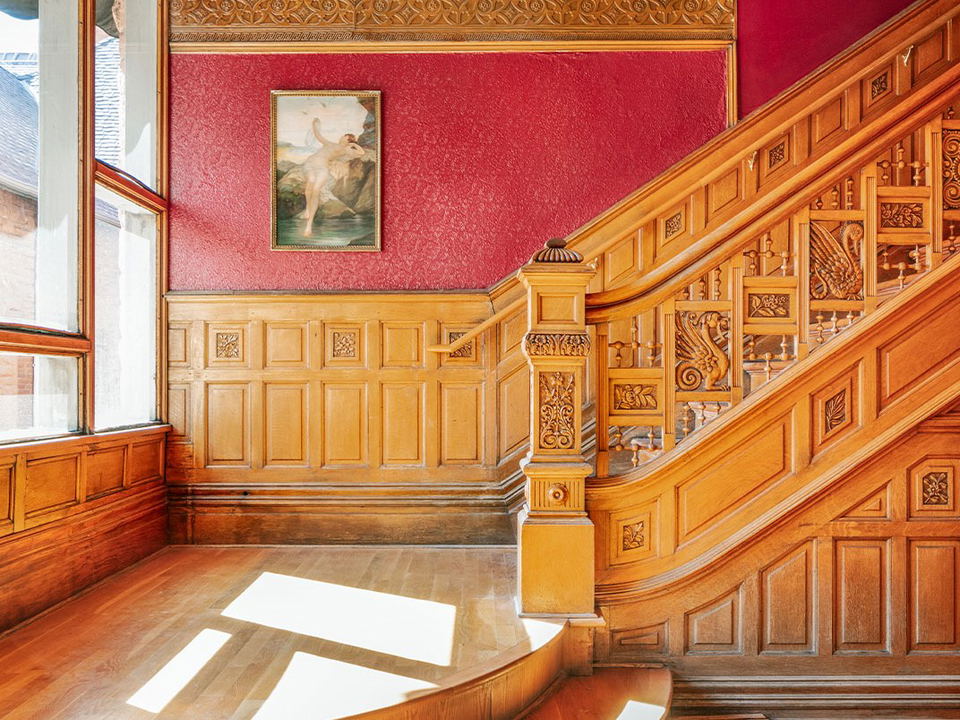 An ornate Victorian-era wooden staircase hall with intricate carved balustrades, red walls, coffered ceiling, and wood paneling.
