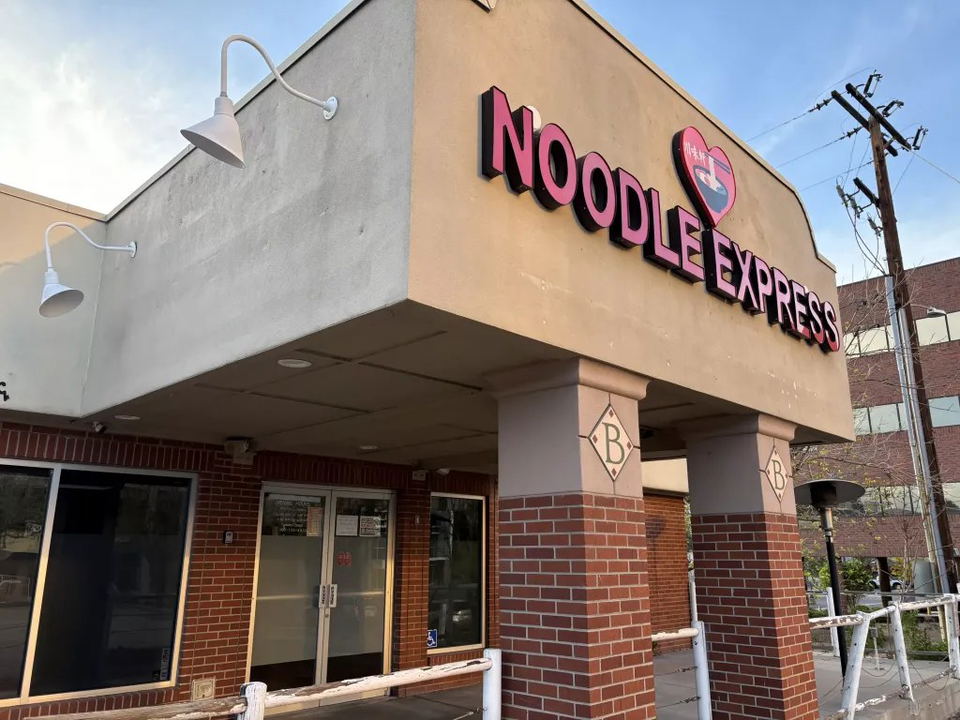 The exterior of Noodle Express at the Belcaro Shopping Center, with its pink and red sign visible on the facade.