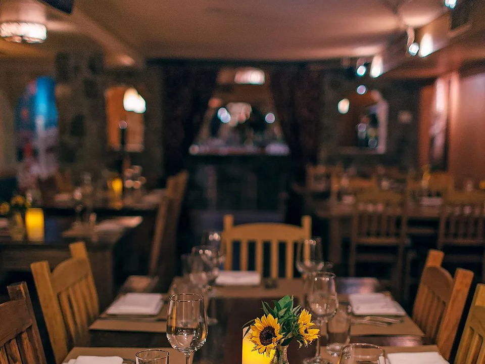 A warmly lit Italian restaurant dining room with wooden tables set with glassware, white napkins, sunflower centerpieces, and candles.