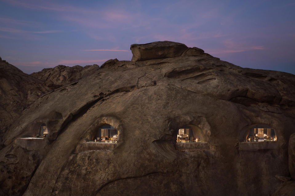Cave-style hotel rooms carved into desert rock formations, illuminated at twilight, photographed from exterior.