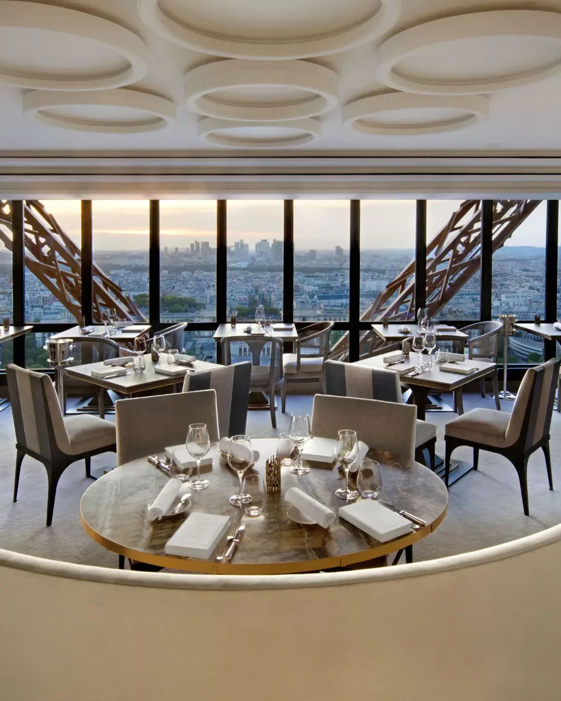 An elegant dining room with tables set for dinner, offering panoramic views of the Paris skyline and the Eiffel Tower's structure.