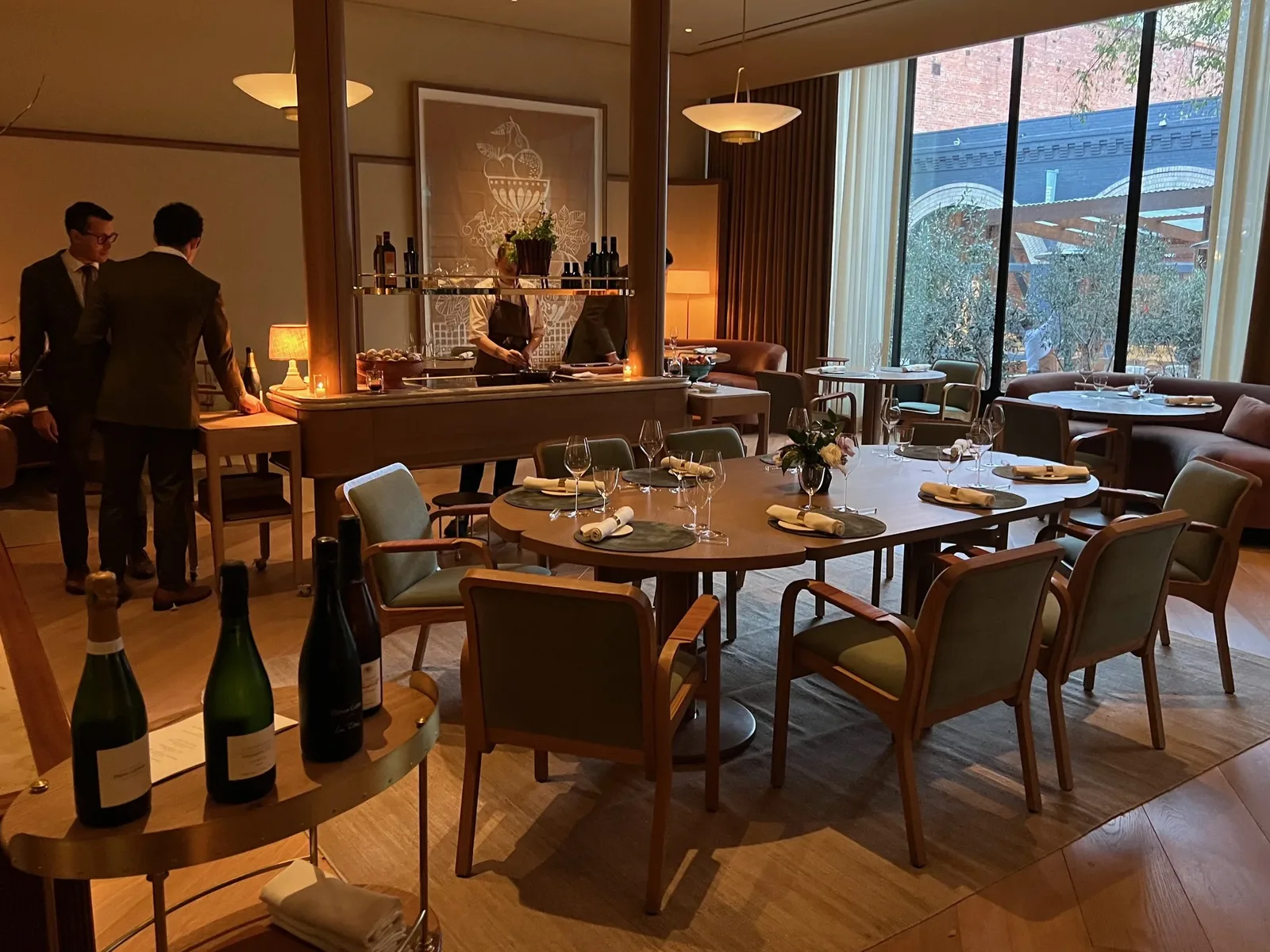 An upscale restaurant interior with round dining tables, wooden chairs with green cushions, wine bottles, and large windows showing an urban view.