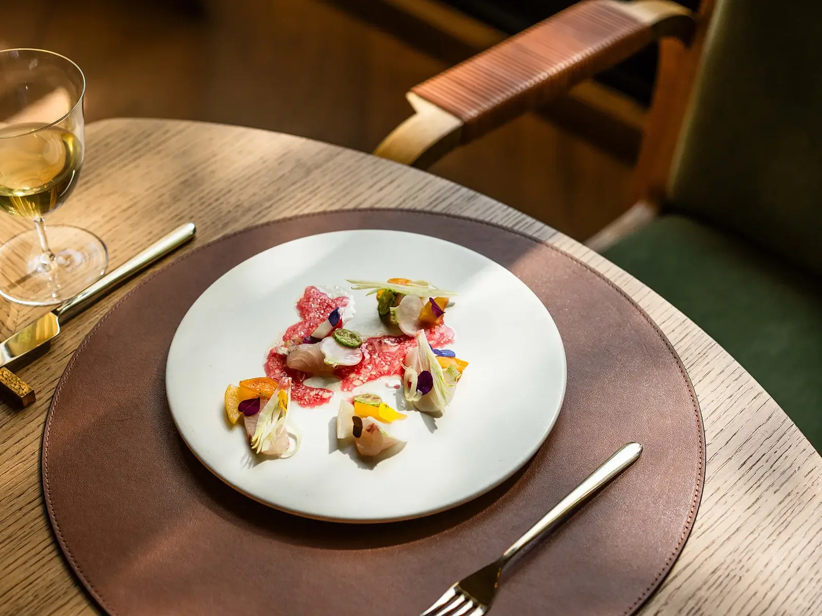 A white plate with an artistic arrangement of sliced cured meat, colorful edible flowers, and vegetable garnishes, placed on a brown leather charger at a wooden table with a wine glass nearby.