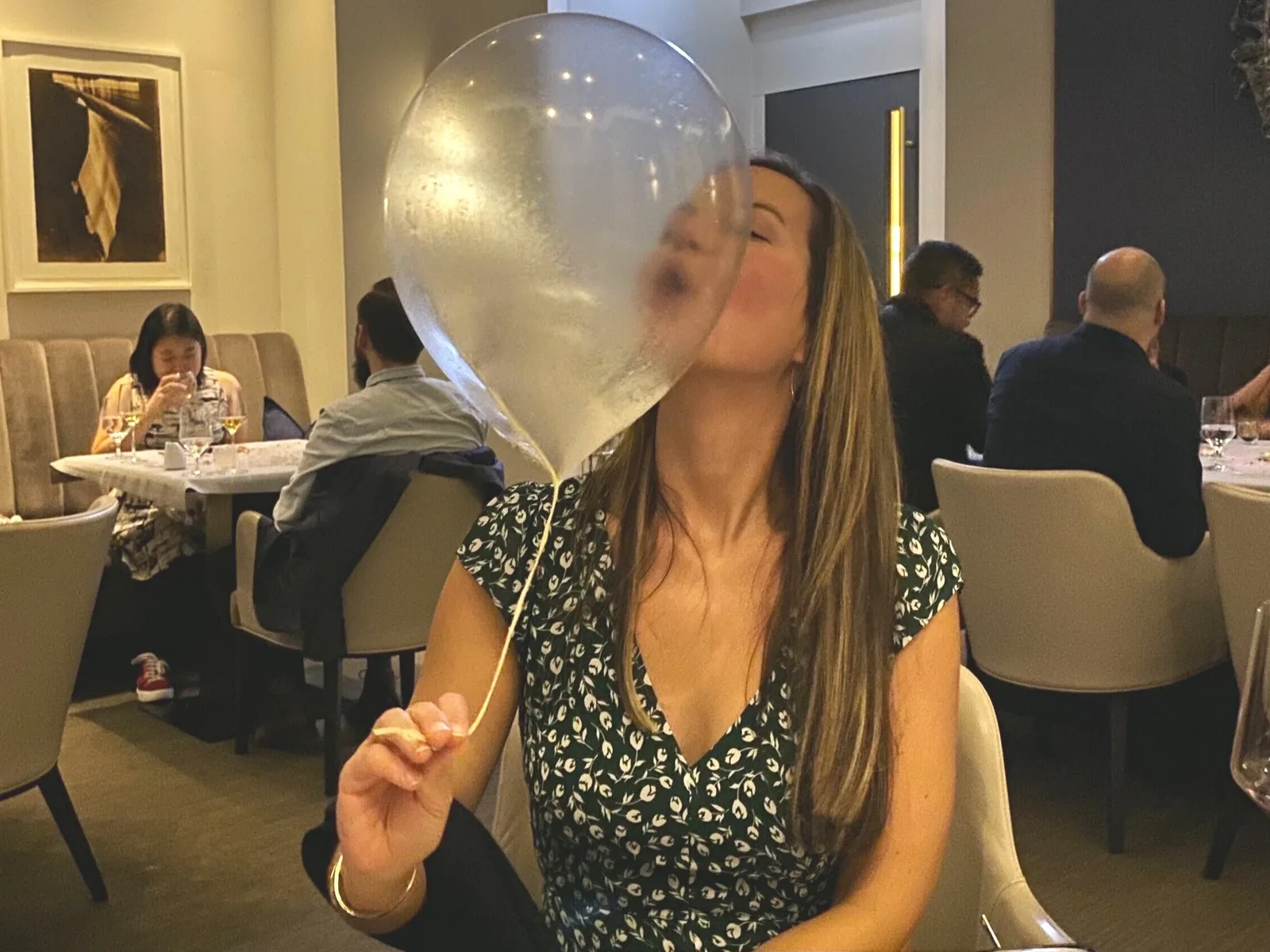 A woman in a dark green floral dress blows into a clear balloon, creating a large bubble, at Alinea Restaurant.