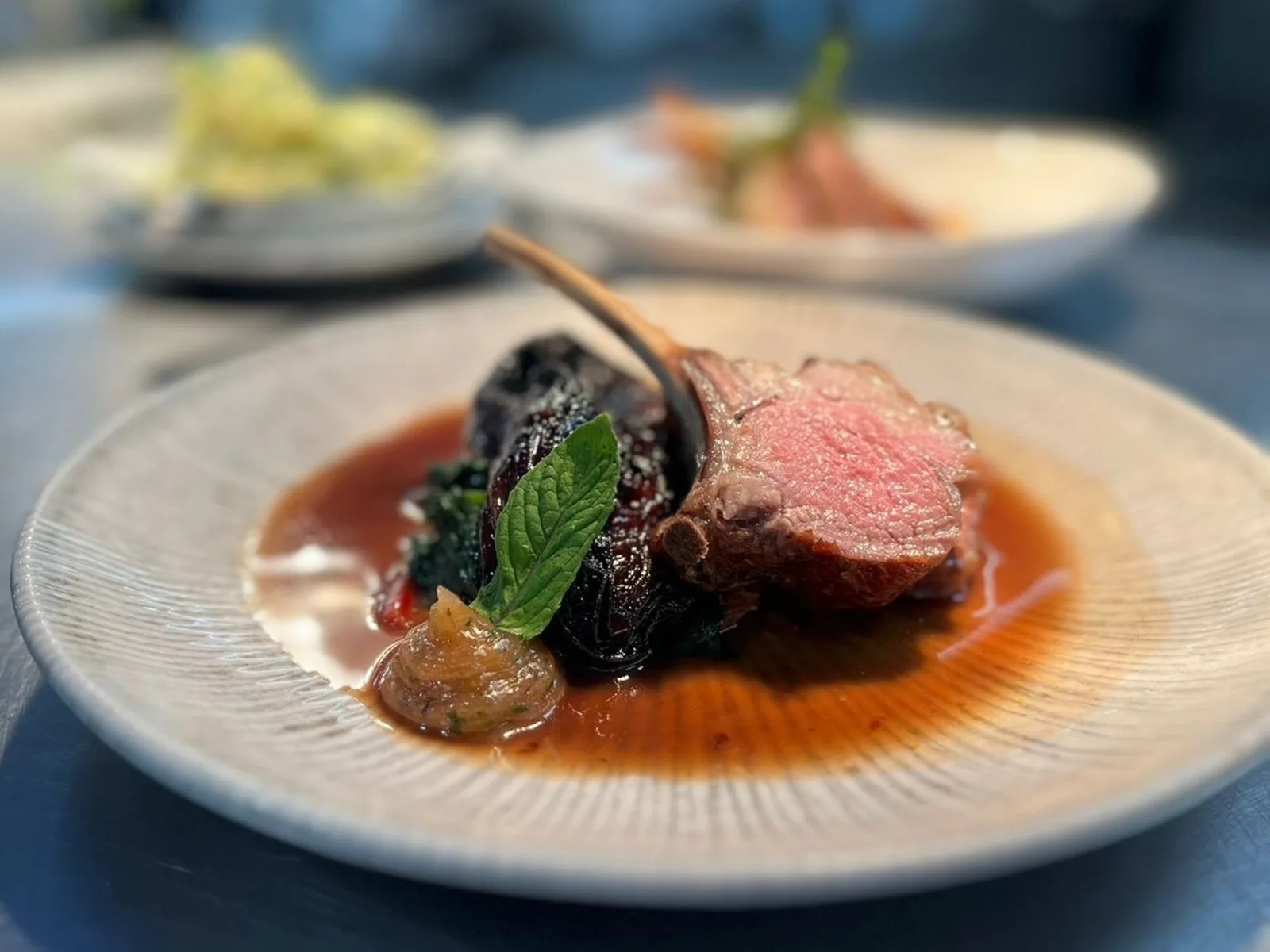 A white plate with seared lamb, dark roasted beets, sauce, and mint leaf garnish, with additional plated dishes visible in the soft-focused background.