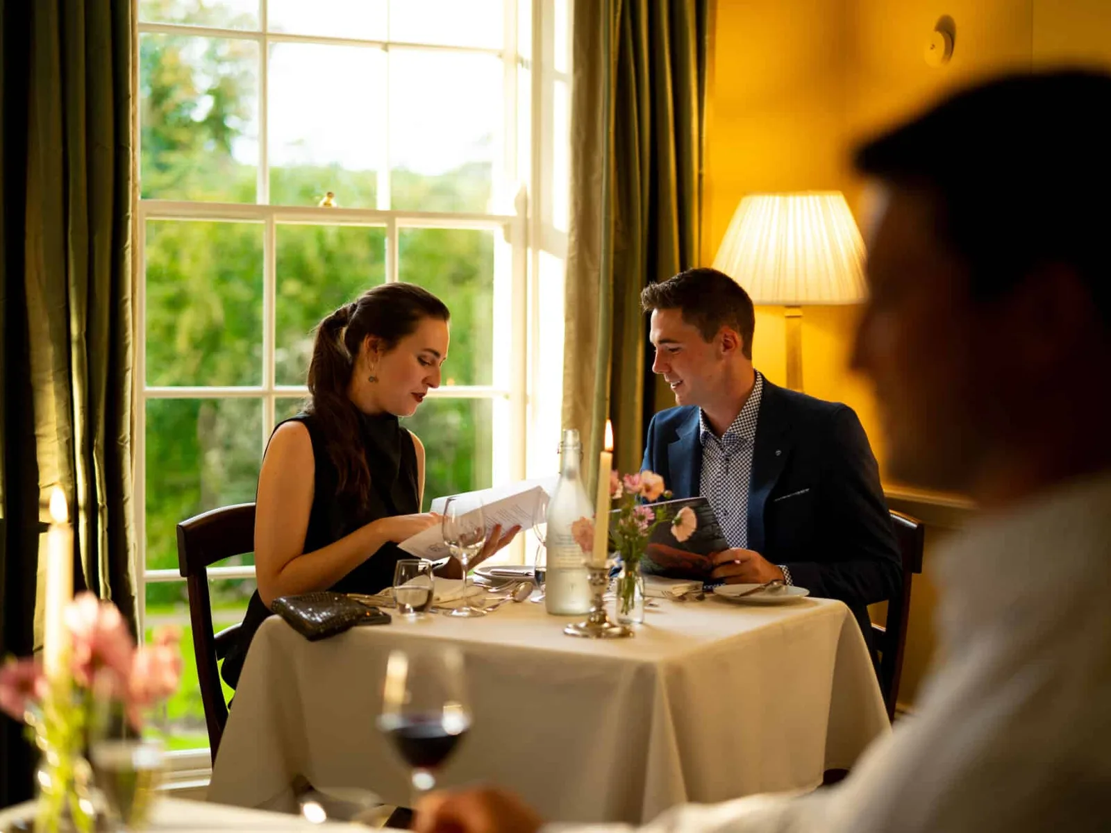 Two people dining at a table with white tablecloth in a bright yellow room with tall windows and garden views.