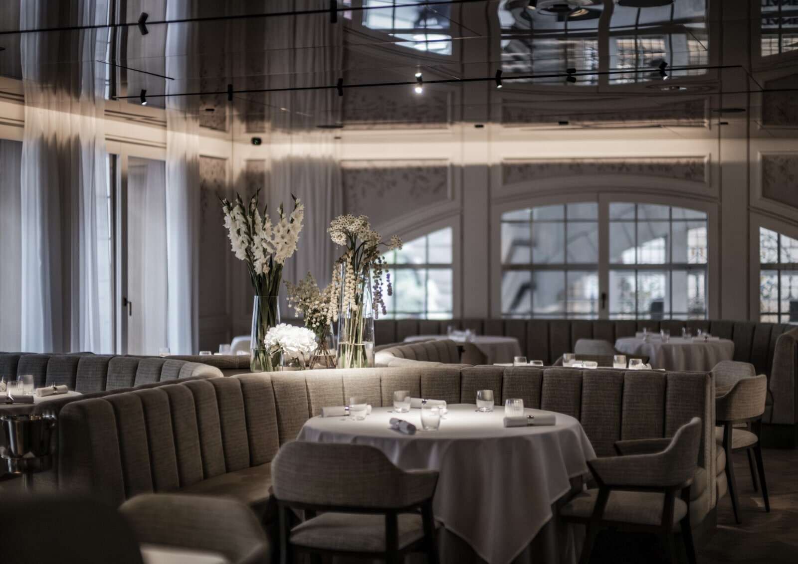 A luxurious dining room with plush, curved banquettes, round tables set with white tablecloths, and tall vases of white flowers.