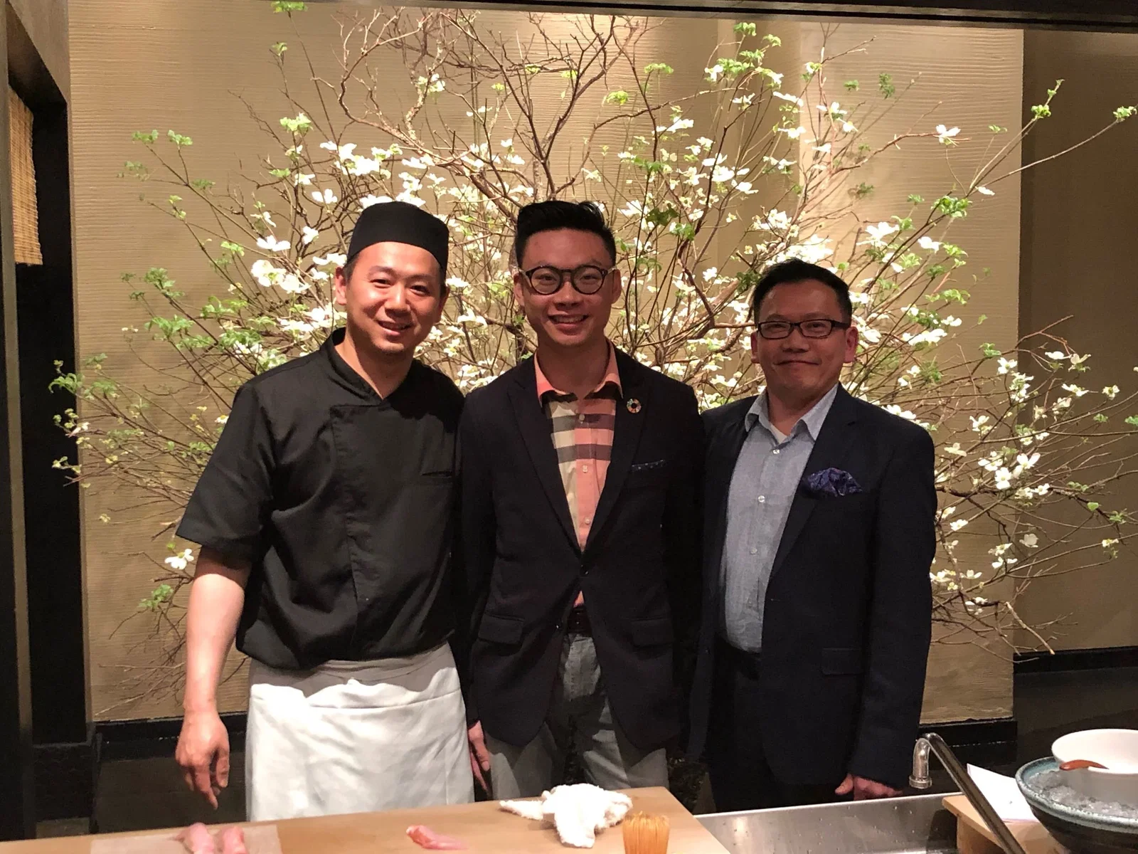 Three men, one in a chef's uniform and two in suits, stand smiling in front of a wall adorned with white flowering branches.