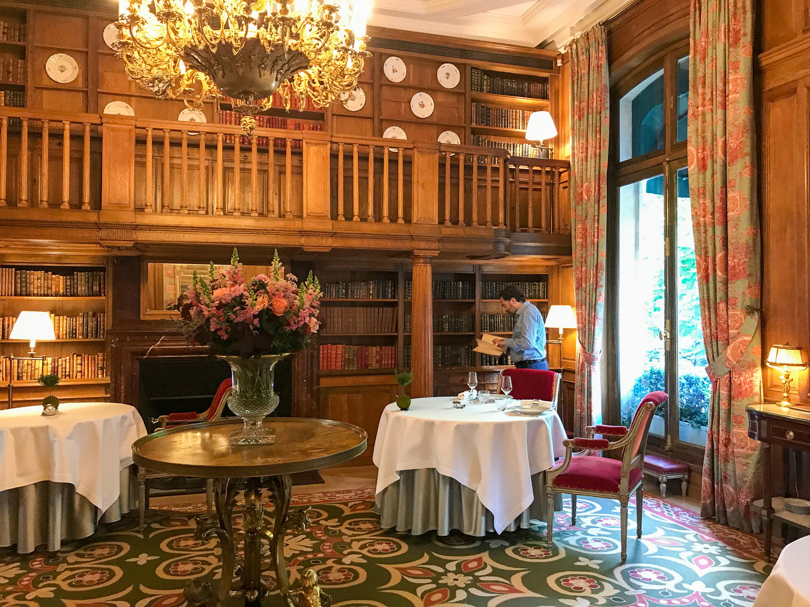 A luxurious dining room with wood-paneled walls, bookshelves, and a grand chandelier. Tables are set with white tablecloths, and a man is seen