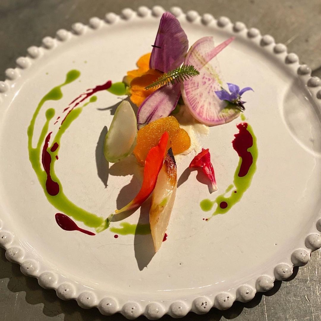 A white plate holds a colorful, artfully arranged dish with sliced radishes, orange segments, and green and red sauces.
