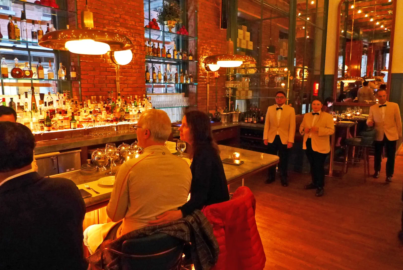 Diners seated at a long bar, with a wall of liquor bottles behind them and servers in beige jackets nearby.