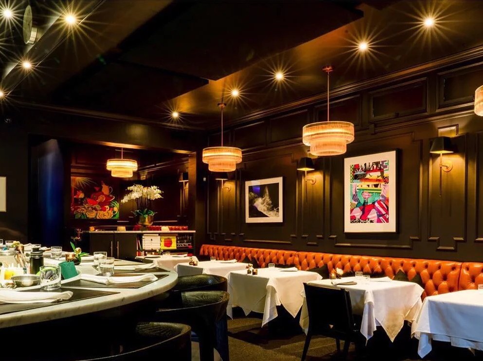 An upscale restaurant interior with dark walls, white-clothed tables, orange tufted banquette seating, hanging chandeliers, and colorful framed art.