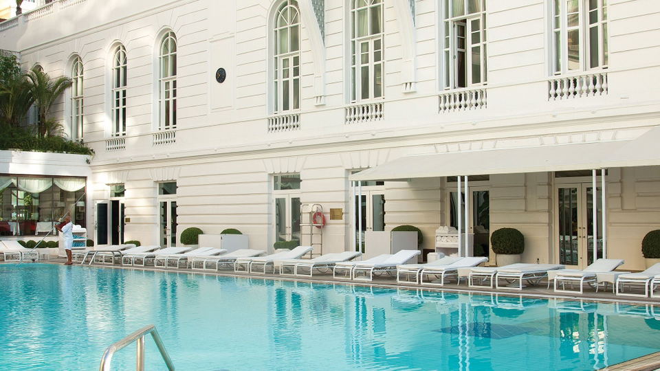 A luxury hotel pool area with white neoclassical architecture, arched windows, balustrades, white sun loungers, and turquoise pool water.
