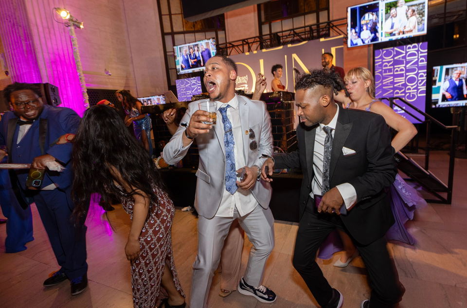 People in formal attire dance at a lively event, with large screens displaying images and text in the background.