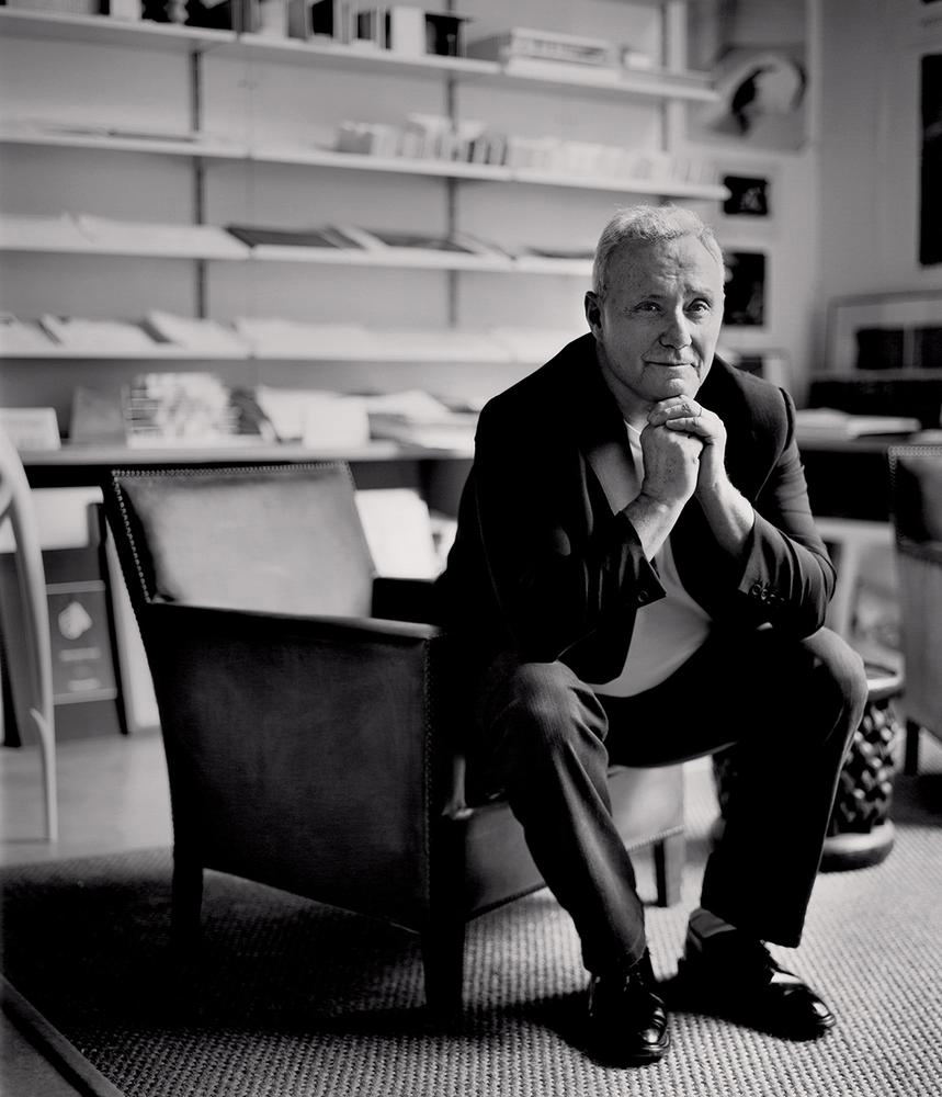 A black-and-white editorial portrait of Ian Schrager, an older man with gray hair, seated in an armchair with his hands clasped.