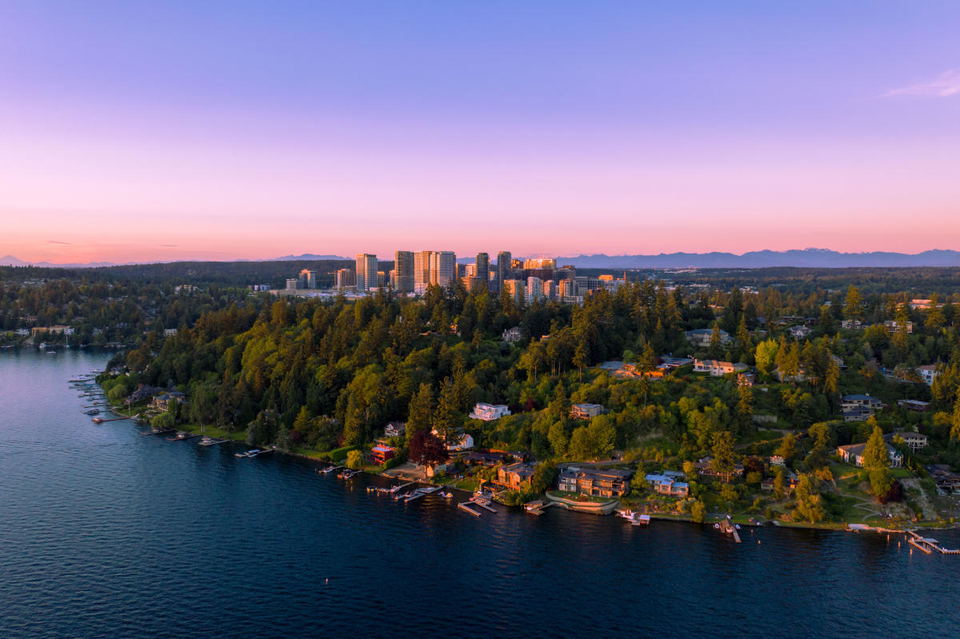 Aerial view of Downtown Bellevue skyline with high-rise buildings, surrounded by dense green forests and luxury homes along a lakefront.