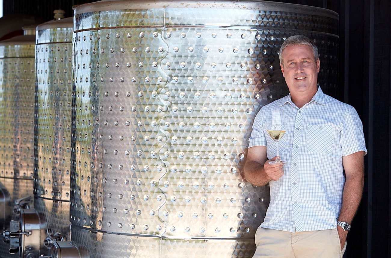 Adam Lowy, founder of Cloudsley Cellars, stands smiling in a winery cellar, holding a glass of white wine next to large stainless steel fermentation