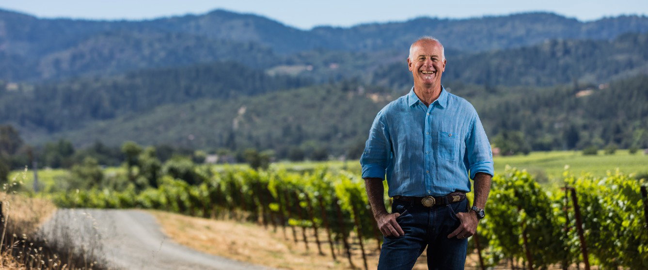 A smiling middle-aged man in a blue linen shirt stands confidently in front of vineyard rows with mountains in the background.