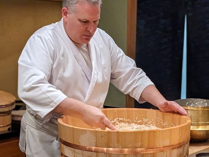 A white-uniformed male chef leans over a large round wooden hangiri tub, hands spread across seasoned sushi rice, in a dimly lit Japanese restaurant kitchen.
