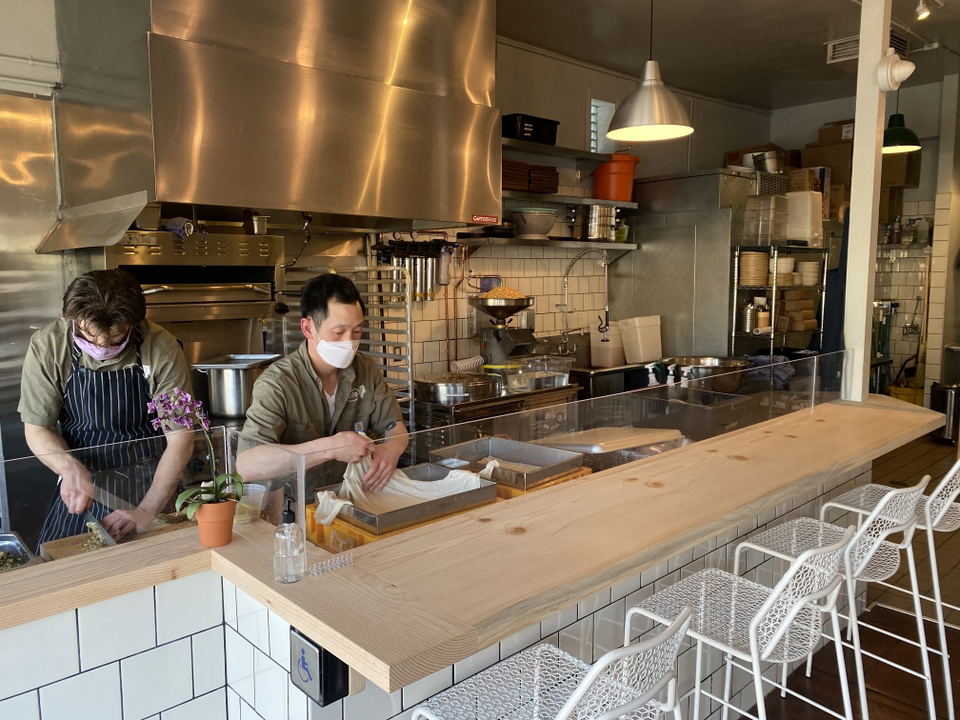 Two masked chefs prepare food behind a counter in an open kitchen, with stainless steel appliances and white tile walls.
